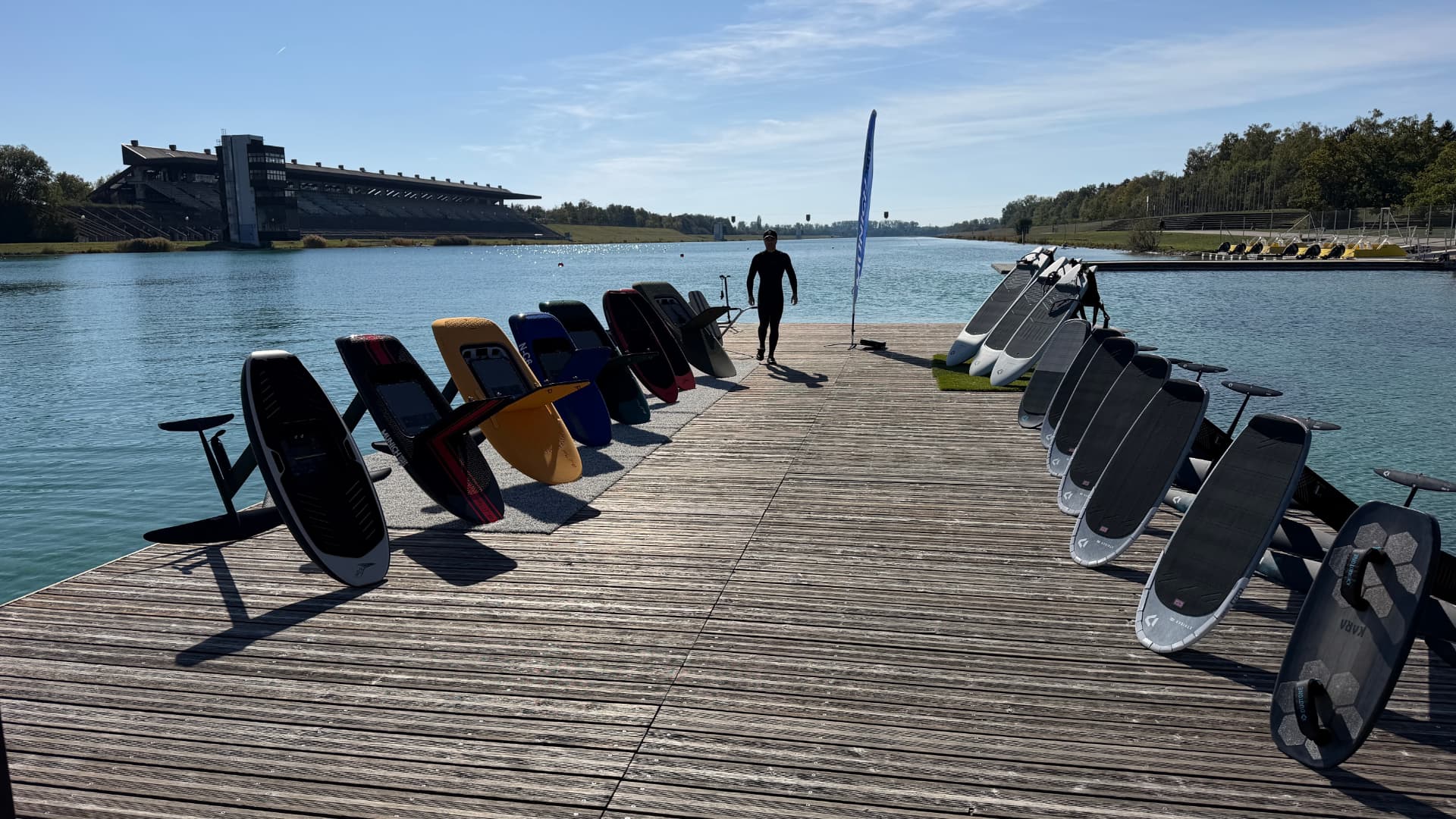 Foilboards auf dem Holzsteg an der Olympia-Regattaanlage München, eine Person im Neoprenanzug läuft auf die Kamera zu